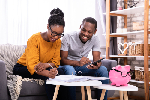 Smiling woman and man sitting on a couch, woman writing in folder on a coffee table, man with calculator in hand, pink piggy bank with glasses on also on coffee table