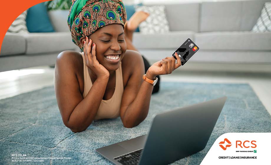 Smiling woman laying on a carpet, holding a RCS credit card, busy on her laptop.